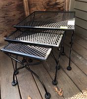 Photo showing three black painted wrought iron nesting tables stacked together on a wood porch floor, highlighting the mesh tops and legs.