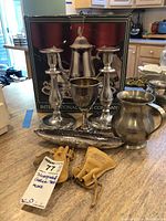 Silverplated coffee set components displayed with candle holders, mug, and napkin rings on countertop with box in background.