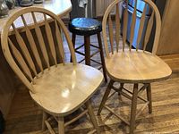 Two light-colored wooden swivel stools with spindle backs and a smaller wooden stool with a round repaired vinyl top arranged on a hardwood floor in a kitchen setting.