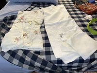 Photo showing two white tablecloths, one with colorful floral embroidery along the border and cutwork, and one plain with subtle embroidery.