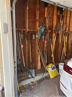 Wide view of lawn and garden tools hanging on a wooden wall in a garage including rakes, shovels, hoes, and pruners.