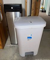 Photo showing both trash bins side by side; white plastic StepOn pedal bin in front, taller stainless steel touchless bin behind.