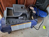 Full side view of blue Campbell Hausfeld air compressor with black top housing, dual gauges and blue coiled air hose on concrete floor.