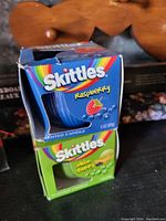 Two boxed Skittles brand scented candles, Raspberry (blue) and Melon Berry (green), both 3 oz size.