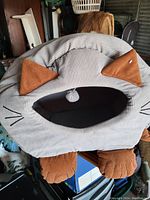 Photo of a corduroy fabric cat house shaped as a gray cat face with brown ears and paws, featuring an open circular entrance as the mouth.