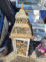 Photo of the decorative metal lantern showing its ornate top, lattice cutouts, and floral metalwork on glass panels, placed among other assorted items.