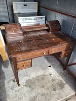 Front and top view of a dark wooden desk with multiple drawers and two raised compartments on top, showing wear and surface scratches.