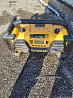 Yellow DeWalt radio with black metal protective frame placed outdoors on asphalt, showing control panel and speakers on front.