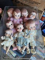 Photo showing eight vintage baby dolls lying in a clear container, each wearing different knitted or fabric clothing.