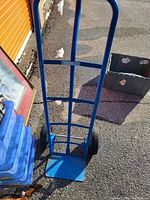 Blue metal dolly hand truck standing upright on an asphalt surface outdoors, with a plastic crate and plastic storage bins nearby.
