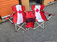 Two connected folding chairs featuring red and white fabric with a Canadian maple leaf design, white metal frames, armrests with cup holders, and central fabric utility compartment.