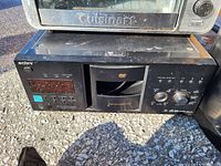 Photo showing a Sony DVD Disc Play Center model DVP-CX995V in black with buttons, dial knobs and disc tray. Above it sits a stainless steel Cuisinart toaster oven with visible wear.