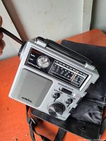 Front angled photo of silver and black portable emergency radio showing speaker, analog frequency dial, flashlight, tuning controls, and LED indicators.