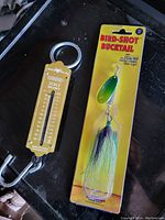 Photo showing a yellow metal scale balance and a packaged green and yellow Bird-Shot Bucktail lure with feather tail.