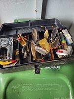 Open black fishing tackle box showing various metallic fishing lures including spoons and spinnerbaits with colorful beads and hooks.