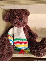 Photo of brown Hudson Bay plush bear wearing a multi-colored striped scarf with tassels, sitting against a beige wall.