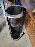 Image of black Keurig coffee maker with silver top accent, showing front view with drip tray and power cord on floor.