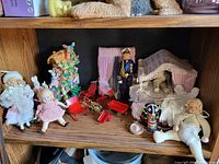 Wide shot of wooden shelf containing various dolls, a police officer figurine, miniature rocking chairs, colorful bird figurine, baby cradle with bedding, and small accessories.