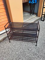 Photo showing a three-tier metal shoe rack outdoors on asphalt. The rack is dark brown with horizontal bars and side supports.
