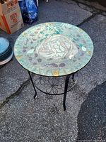Photo of the round side table outdoors showing mosaic glass top with colorful design and black metal frame with decorative elements.