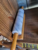Marble rolling pin resting on a wooden display stand. The rolling pin is white with grey veins and has wooden handles on both ends. The wooden stand is made from a light polished wood and supports the pin horizontally.