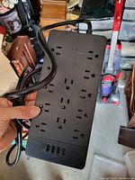 Photo of a black surge protect power bar held by a hand showing six AC outlets and four USB charging ports with power cord attached