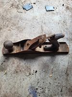 Photo of a single vintage metal hand wood plane with wooden front knob and rear handle, placed on a rough surface showing rust and wear.