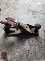 Single hand plane tool placed on a concrete surface, showing metal body and wooden handles with visible rust and wear.