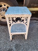 Front view of a light-colored wicker side table showing woven top, shelf, and decorative sides.
