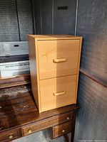 Wooden file cabinet with two drawers and wooden handles, placed on wooden desk with kitchen appliances in background