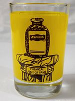 Front view of vintage small glass with yellow background and black aspirin bottle design on Chemical Hall of Fame pedestal.