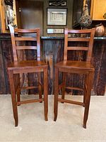 Front view of two cherry stained wooden barstools with ladder backs, showing wear and surface marks from age and use.
