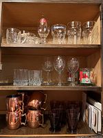 Shelf with multiple glassware items categorically arranged; beer glasses on top two shelves, copper mugs and purple glasses on bottom shelf.