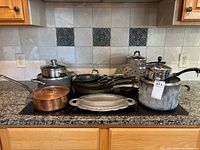 Overview of the entire cookware lot including copper pot, Calphalon saucepan, cast iron skillets, and other metal pots and pans on kitchen countertop stovetop.