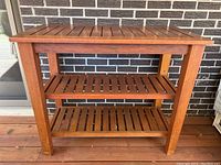 Front view of teak buffet table against a brick wall, showing slatted top and two lower shelves.
