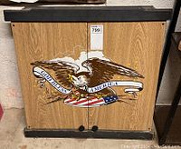 Closed view of wooden dartboard cabinet with front patriotic graphic showing eagle and American flag with text 'God Bless America'.