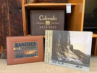 Three books shown together on the floor beside wooden furniture: large brown hardcover 'Colorado 1870-2000', smaller leather-bound 'Ranches of Colorado', and the Curtis book with image of cliff and riders on horses on cover.