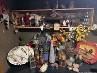 Photo showing a wooden shelf with assorted Christmas and folk art decorations including bird and squirrel candles, metal rooster, faux flowers, decorative plates, small lanterns, and artificial apples in a bowl.