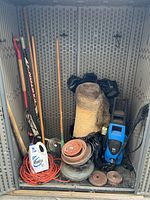 Photo showing garden tools including shovel, wooden handles, extension cord, detergent jug, rolls of plastic or fabric, and a blue/black electric pressure washer.