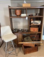 Wide view showing bar stool, media cabinet with shelves, Danish serving pieces on shelves, wooden crates stacked on floor and inside cabinet, model car box on shelf, and a wooden bowl on top shelf.