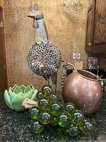 Photo showing all four items: metal rooster sculpture, green lotus candle holder, Lucite grape cluster, and copper wine bottle holder on a granite countertop.