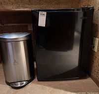 Black mini refrigerator and silver Simplehuman trash can standing side by side on a tiled floor against a wooden cabinet.