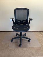 Front view of black mesh office chair positioned on clear plastic floor mat, showing mesh seat and backrest, armrests with visible wear, and five-wheel base.