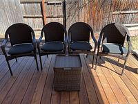 Four plastic wicker patio chairs with black cushions and a matching plastic wicker storage box side table arranged on a wooden deck under sunlight.