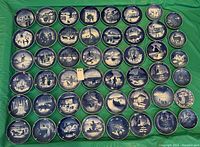 Top view showing the full collection of 48 Royal Copenhagen blue and white collector plates arranged flat on a green surface, displaying diverse winter holiday scenes.