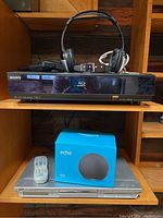 Sony Blu-ray player on top shelf with black Bluetooth headphones and small device on top, JVC DVD player with remote and boxed Amazon Echo speaker below.