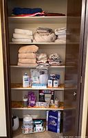 View of closet shelves with stacked towels, folded sheets, linens, and assorted toiletry items including lotion, bath salts, toothpaste tablets, and tissue box.