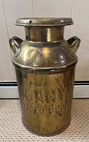 Front side view of vintage metal milk jug can showing two handles and lid on top, placed on carpet with white wooden panel background.