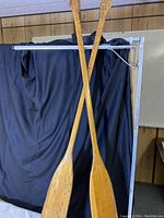 Two aged wooden paddles crossed in front of a black background and wood panel wall, showing full length and surface wear.
