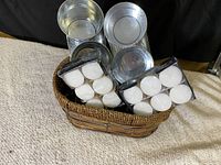 Woven basket with metal candle holders and multiple packs of white tealight candles inside, arranged to show contents.
